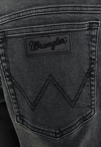 Jeans de mezclilla negra con un bolsillo trasero que lleva una etiqueta rectangular de cuero con la inscripción "Wrangler". La costura forma un patrón distintivo en forma de W.