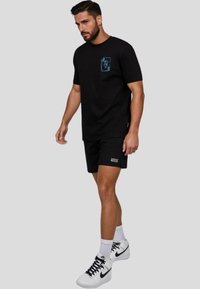 Černé bavlněné tričko s tyrkysovým grafickým detailem na kapse, spárované s černými šortkami, bílými ponožkami a bílými teniskami Nike.