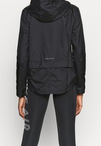 Veste légère noire avec une capuche, tissu texturé, taille cintrée et accents réfléchissants le long de l'ourlet. Associée à des leggings noirs.