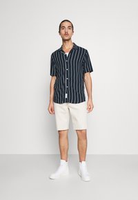 Dunkelblaues Kurzarmhemd mit vertikalen weißen Streifen, kombiniert mit beigefarbenen Shorts und weißen High-Top-Sneakers. Einfaches Design mit durchgehend geknöpftem Vorderteil.