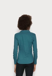 Camicia a maniche lunghe color teal con colletto, caratterizzata da un design aderente e polsini con bottoni; tessuto liscio con una leggera lucentezza. Visibili pantaloni neri.