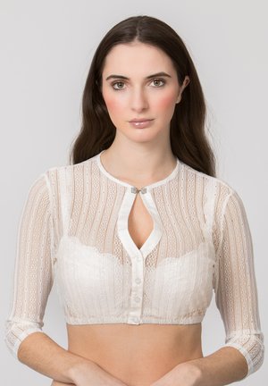 Young woman with long brown hair wearing a white sheer lace cropped cardigan with buttons and a keyhole neckline, standing against a plain background.