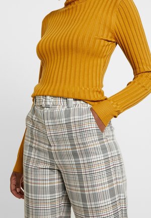 Maglione a collo alto a righe senape abbinato a pantaloni a vita alta a quadri con motivi a griglia nera, bianca e rossa. Tessuto testurizzato.