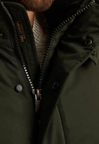 Zelená parka s vysokým límcem, zapínáním na zip a patentkami. Obsahuje texturovaný béžový svetr pod ní a štítek s logem na zipu.