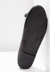 Semelle d'une ballerine grise avec des motifs en relief de chaussures de ballerine et la marque "Pretty Ballerinas", reposant contre un coin blanc.
