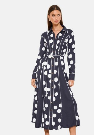 Žena v tmavě modrých midi šatech s velkými bílými puntíky, vertikálními bílými pruhy, páskem v pase a hnědými knoflíky na přední straně.