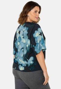 Svart topp med blå blommönster, korta ärmar och avslappnad passform. Bärs med grå byxor, som visar en slät tygtextur.