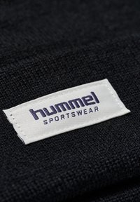 Tissu noir avec une surface texturée, comportant une étiquette tissée blanche avec "hummel SPORTSWEAR" en texte violet. L'étiquette est solidement cousue.