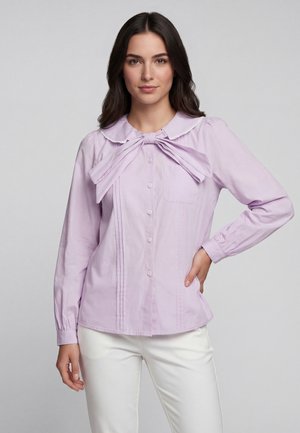 Frau trägt eine hellviolette Bluse mit großer Schleife und weiße Hose, steht mit einer Hand auf der Hüfte vor einem einfarbigen Hintergrund.