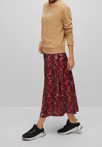 Sudadera marrón con texto blanco, falda plisada con estampado rojo y zapatillas negras con suelas blancas y marrones. Atuendo casual en un entorno neutro.