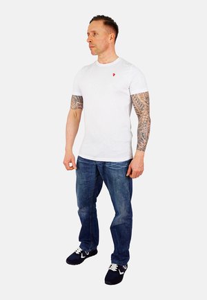 Tricou alb din bumbac cu guler rotund și un mic logo roșu pe piept, purtat împreună cu blugi albaștri din denim și adidași navy cu șireți albi.