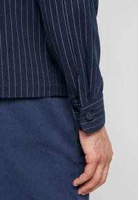 Camisa a rayas azul marino con finas rayas blancas, con puño abotonado. La tela tiene un acabado texturizado, mostrada junto a pantalones azul marino.