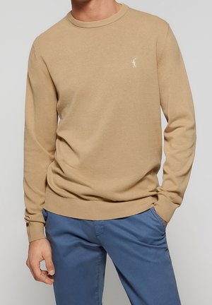 Uomo che indossa un maglione beige a maniche lunghe con un piccolo logo bianco di un giocatore di polo e pantaloni blu, in piedi con una mano in tasca.