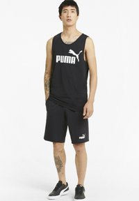 Canotta nera senza maniche con logo bianco "PUMA", pantaloni corti neri abbinati e scarpe nere con dettagli bianchi. Tatuaggi visibili sulle braccia.