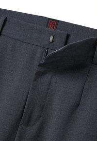 Pantalons taillés gris foncé en tissu texturé, avec une ceinture lisse, une fermeture éclair en métal et une étiquette de marque rouge à l'intérieur.