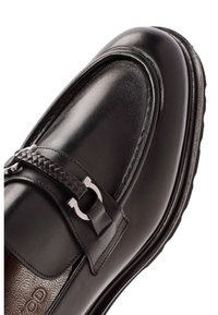 Mocasín de cuero negro con una tira de acento trenzada y detalle de hebilla metálica. El zapato tiene una textura suave y una suela resistente.