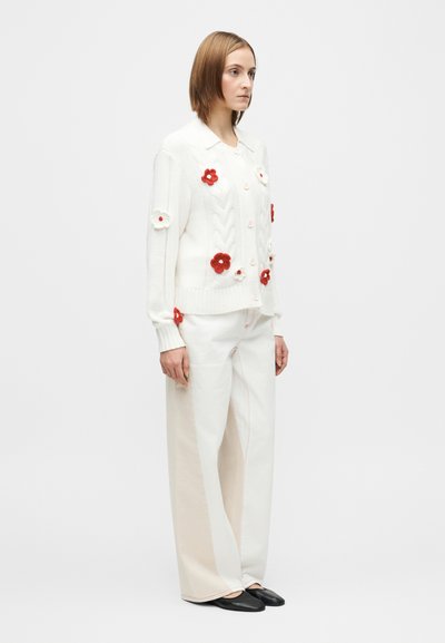 Hvit strikket cardigan med røde og hvite blomsterapplikasjoner, kombinert med brede kremfargede bukser med røde sømmer og svarte sko.