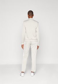 Ανοιχτό γκρι σετ φόρμας κατασκευασμένο από λείο ύφασμα, με λευκές ρίγες στα πλάγια. Διαθέτει στρογγυλή λαιμόκοψη και ελαστικές μανσέτες. Οι αθλητικές εξωτερικές μύτες ολοκληρώνουν την εμφάνιση.