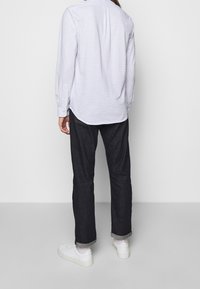 Osoba v bílém vzorovaném tričku s dlouhým rukávem, tmavých vyhrnutých džínách, bílých ponožkách a bílých teniskách stojící zády k jednofonnímu pozadí.