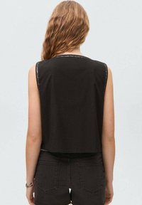 Zwarte mouwloze crop top met een gladde textuur en een gedetailleerde gestikte halslijn. Draagt met donkere denimshorts, wat een casual uitstraling benadrukt.