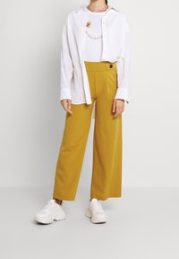 Bílá oversize košile, grafické tričko s květinovým vzorem a široké hořčicové kalhoty. V kombinaci s bílými teniskami. Jednoduchý a neformální design.