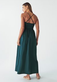 Maxi abito verde scuro con corpetto arricciato, spalline incrociate regolabili e gonna fluida, abbinato a sandali flat.