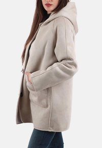 Cappotto beige con cappuccio, dalla texture morbida, con tasche laterali e dettagli delle cuciture. Il design presenta una vestibilità rilassata e un taglio di lunghezza media.