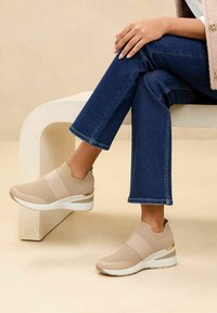 Beige kötött slip-on cipők elasztikus pánttal, texturált felső résszel és fehér wedges talppal, arany díszítéssel. Kék farmernadrággal viselve.
