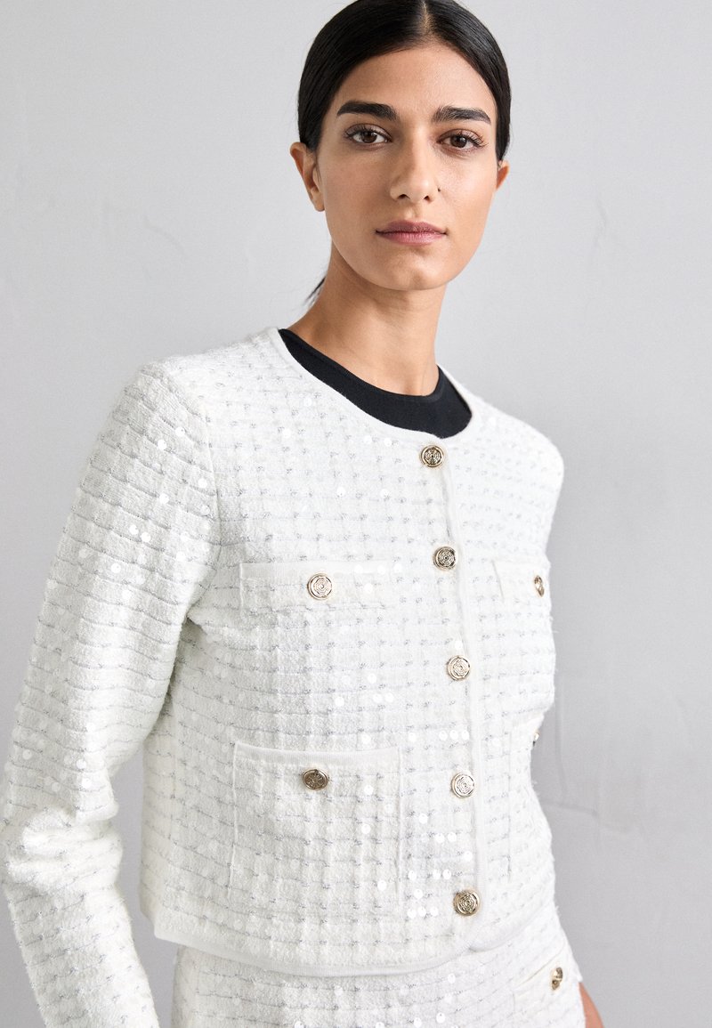 Veste blanche texturée avec un motif matelassé, boutons dorés et deux poches avant. L'encolure est ronde et possède une doublure intérieure noire contrastante.