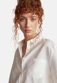 Camicia bianca con bottoni, caratterizzata da un motivo testurizzato, dotata di taschino sul petto e colletto arrotondato. Tessuto morbido e dettagli discreti.