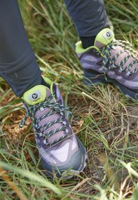 Chaussures de randonnée violettes avec panneaux en mesh, accents verts et lacets à motifs, posées sur l'herbe, mettant en valeur un design robuste et une semelle texturée.