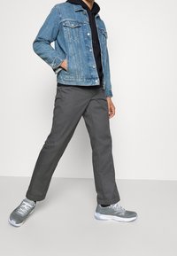 Persona che indossa una giacca di denim blu, un hoodie nero, pantaloni grigi e sneakers grigie, in piedi con le gambe divaricate su uno sfondo chiaro uniforme.