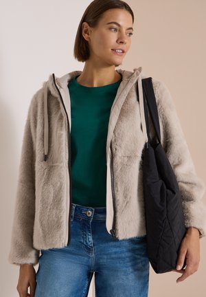 Jeune femme portant une veste en fausse fourrure beige sur une chemise verte et un jean bleu, portant un sac noir matelassé à l'épaule.