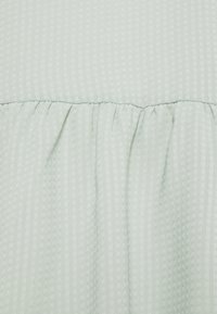 Tissu vert clair avec un motif quadrillé texturé, présentant des détails froncés le long du bord inférieur. Finition douce et lisse.