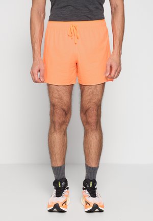 Mann trägt leuchtend orangefarbene Sportshorts, graue Kniestrümpfe und weiße und orangefarbene Laufschuhe, steht vor einem einfarbigen Hintergrund.