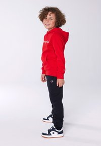 Red hoodie with white lettering, black sweatpants, and black/white sneakers. Features a kangaroo pocket and logo accents. Curly hair.