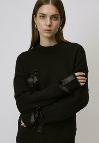 Czarny dzianinowy sweter z wysokim dekoltem, z dekoracyjnymi czarnymi satynowymi wstążkami na rękawach. Miękka tekstura, luźny krój.