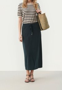 Haut à manches courtes beige et noir à rayures associé à une jupe maxi verte foncée avec une taille à cordon. Sac à main en rotin naturel et sandales noires.