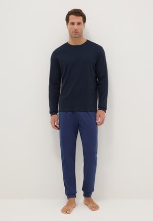 Man staat blootsvoets tegen een effen achtergrond, draagt een marineblauwe lange mouw shirt en blauwe broek, kijkt recht vooruit met een neutrale gezichtsuitdrukking.