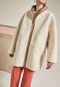 Giacca in pile color crema con zip frontale, collo con accenti marroni e ampie tasche. Il modello indossa pantaloni a coste color ruggine.