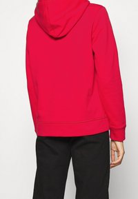 Felpa rossa con vestibilità media, tasca a marsupio e cappuccio a coulisse regolabile. Tessuto morbido e dettagli di cucitura visibili sui polsini. Pantaloni neri.