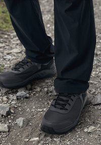 Černé sportovní boty s textilním svrškem, strukturálními panely a gumovými podrážkami. Nosené s tmavými kalhotami na drsném, štěrkovém povrchu.