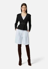 Donna che indossa un blazer nero con cintura, pantaloncini azzurri al ginocchio e stivali marrone scuro sopra il ginocchio, in piedi con le mani nelle tasche.