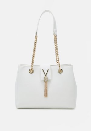 Weiße Handtasche mit goldenen Kettenhenkeln, verziert mit einem goldenen V-förmigen Verschluss und einem hängenden Quasten-Detail vorne.
