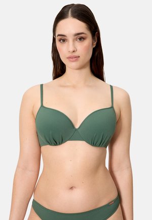 Mujer con un sujetador acolchado verde y bragas de bikini a juego, de pie contra un fondo claro y liso, mirando hacia adelante.