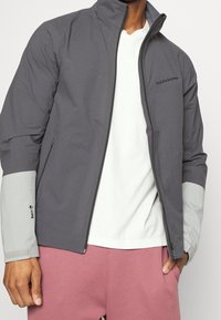 Grijze jas met hoge kraag, grijze accenten op de mouwen, ritssluiting en logodetail. Gecombineerd met een licht shirt en roze korte broek.