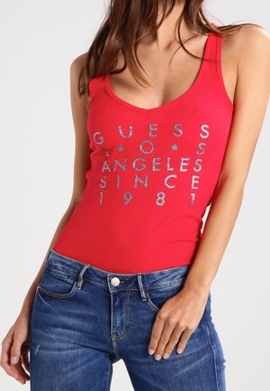 Kvinde iført en rød tank top med sølvfarvet tekst og blå jeans, poserende med den ene hånd hvilende på hendes lår mod en ensfarvet baggrund.