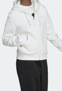 Sweat-shirt zippé blanc avec capuche à cordon, coupe décontractée, poches avant et poignets élastiques. Associé à un pantalon noir. Tissu doux et texturé.