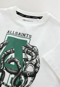 Valge puuvillane t-särk, millel on graafiline kujundus rohelistest tähtedest "A", mis on põimunud musta madu motiiviga. Etikett on näha kaeluses.