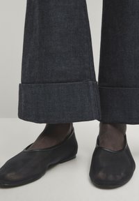 Chaussures à enfiler en mesh noir avec un bout arrondi et une semelle plate, associées à un pantalon en denim foncé à jambes larges avec un ourlet replié.